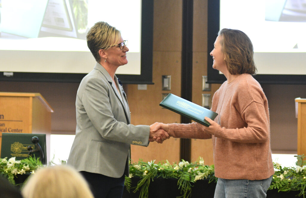 Woman in copper colored sweater receives a hand shake and a green folder from a woman wearing a grey sports jacket. 