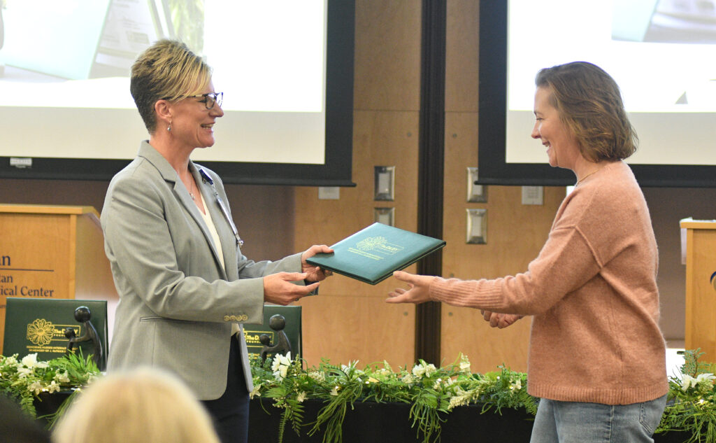 Woman in copper colored sweater receives a green folder from a woman wearing a grey sports jacket. 