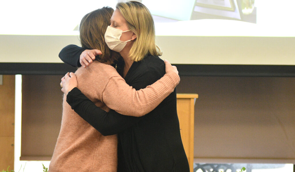 Woman in copper colored sweater hugs a woman wearing a black sweater and a face mask. 