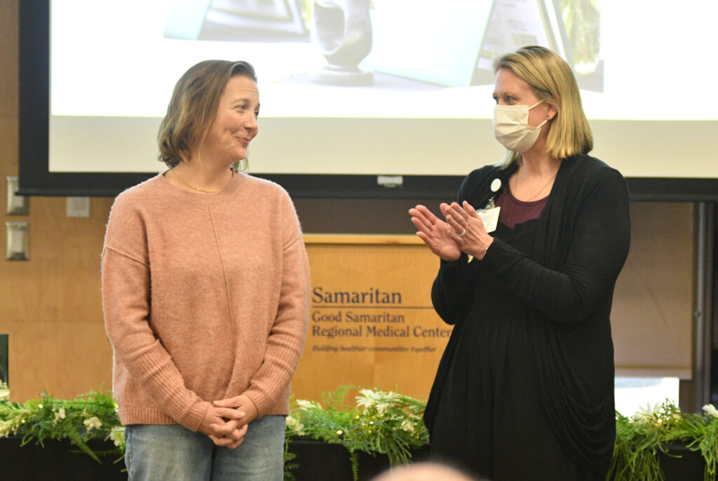 Woman in copper colored sweater stands next to a woman wearing a black sweater and a face mask. 