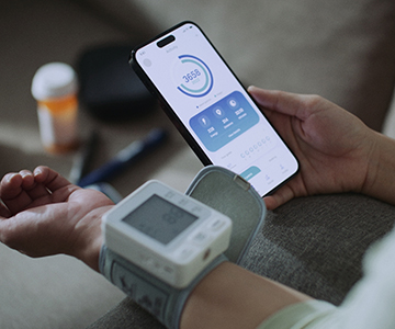 Image with a blood pressure sleeve on wrist, a cell phone in hand and a pill bottle in background demonstrating importance of being on top of your health.