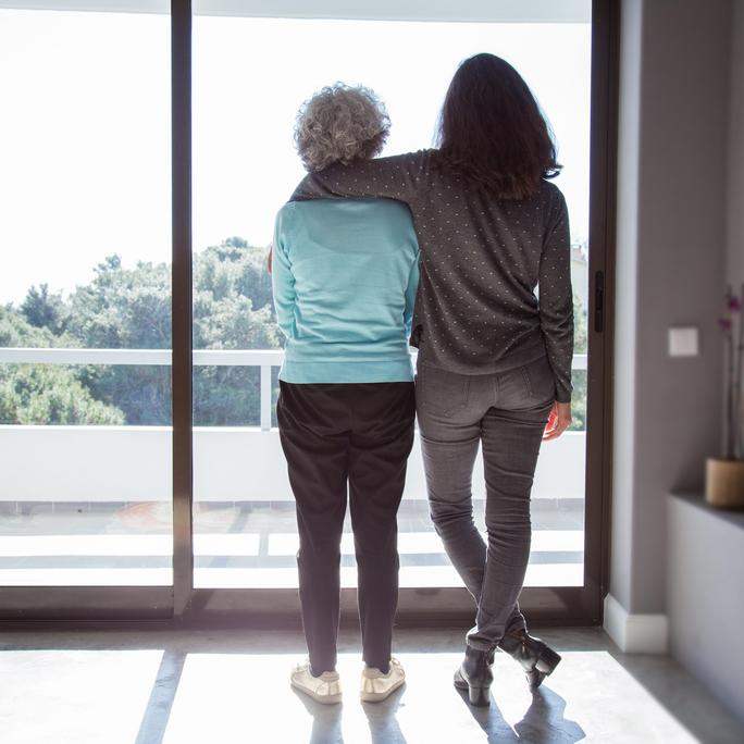 Adult daughter and mother standing and side hugging while looking out the window.
