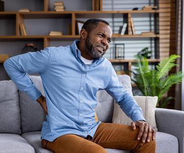 Adult Black make sitting on a sofa, grabbing his lower back and making an expression that indicates pain.
