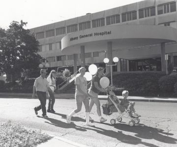 A family walks in front of the Albany General Hospital smiling and carrying balloons.
