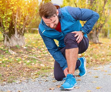 Man with blue jacket on jog path kneeling down touching ankle sports injury