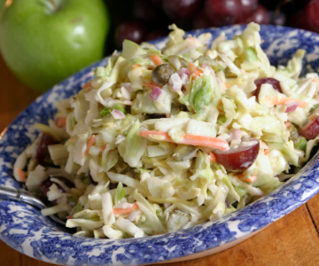 A blue and white patterned dish filled with creamy green apple coleslaw. The slaw includes shredded cabbage, carrot strips, diced green apple, and halved red grapes, topped with a light dressing. A green apple and a cluster of grapes are visible in the background.