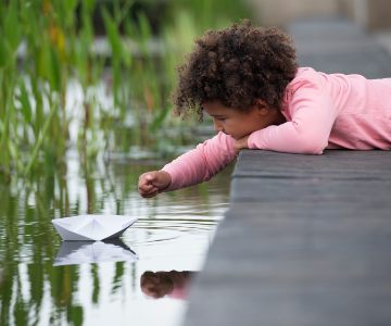 Female child laying on the ground and reaching out to play with an origami piece of art in the pond.