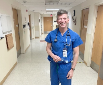 A man in blue scrubs stands in a white hallway with several brown doors.