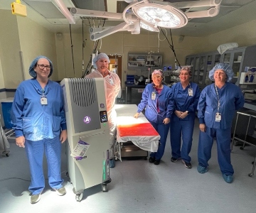 Four women in blue scrubs and one man in white scrubs stand by a operating table and a large grey machine. 