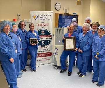 A large group of medical professionals dressed in blue scrubs stand around a vertical award banner in an operating room