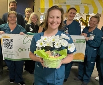 A woman stands with a bouquet of daisies in front of five women and one man standing behind a banner.