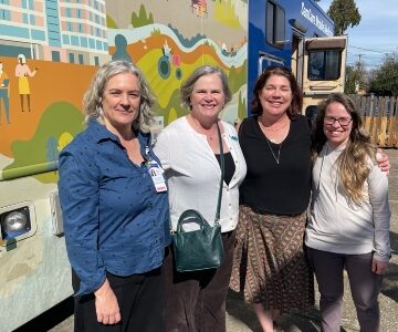 Four women, two in white one wearing blue and another wearing black stand in front of a mural on a mobile medicine unit.