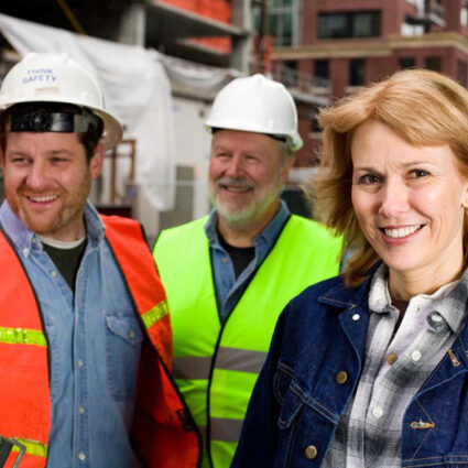 Two male workers with hard hats and visibility vests and a woman in a jean jacket smiling.