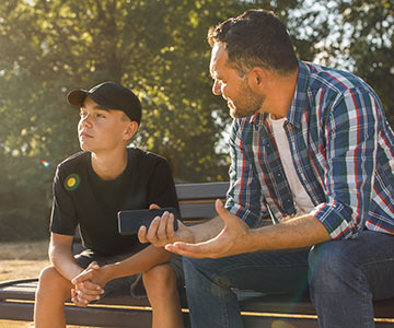 A dad having a heartfelt talk with his preteen son while sitting on a park bench together.