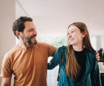 A dad side hugs his daughter as they both look and smile at each other.