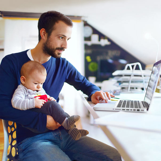 A dad holding a baby while sitting at a desk and using a laptop.