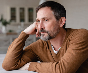 A man sitting in his home resting his head against his hand and looking lost in his thoughts.