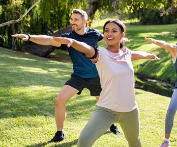 People standing in a grassy park practicing dynamic stretching with their arms spread out and a wide stance.