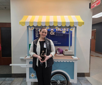 Erika Lentz stands in front of the DAISY Award cart smiling and holding her award.