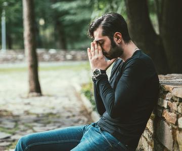 Young adult male sitting in a park with his head in his hands.