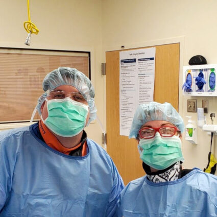 Two people with hairnets, safety glasses, masks and scrubs look toward camera.