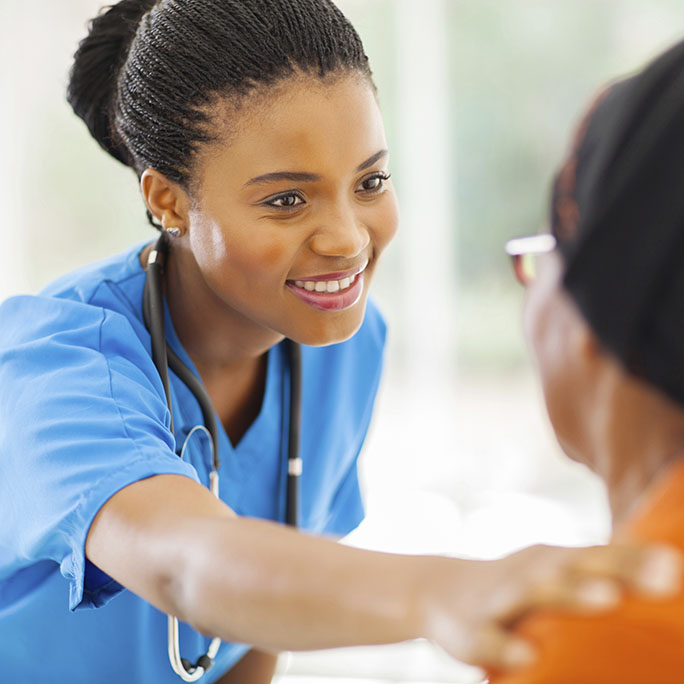 A smiling nurse with a hand on her patient's shoulder in a gesture of comforting care.