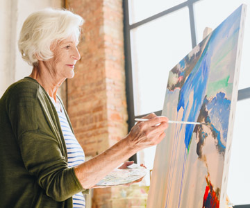 Older woman painting at an easel.