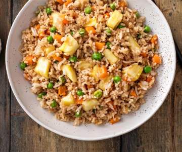 A white bowl sits on a wooden counter and is filled with pineapple fried rice, carrots and green peas.