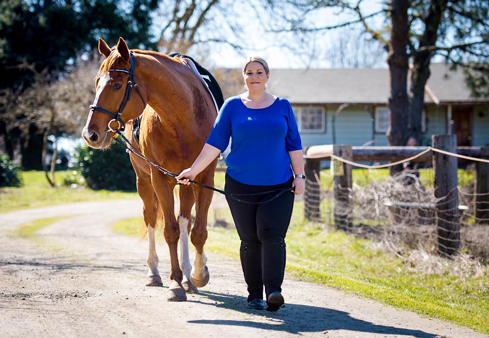 A smiling Amie leading her horse up a gravel lane.