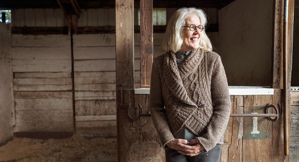 A smiling Suzy standing in the horse barn looking out toward the pasture with her hands folded in front of her.