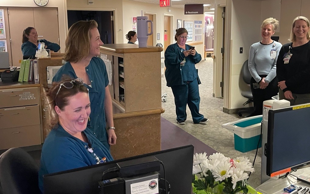 two women wearing nursing scrubs laugh while stand around the nursing desk. 