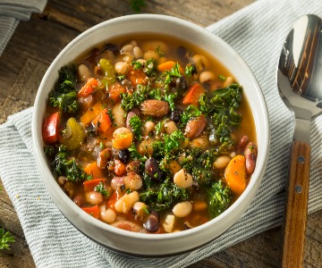 A bowl of hearty vegetable and bean soup, featuring white beans, kidney beans, chopped kale, carrots, celery, and red bell peppers in a clear broth. The bowl is placed on a gray cloth napkin with a wooden-handled spoon beside it.