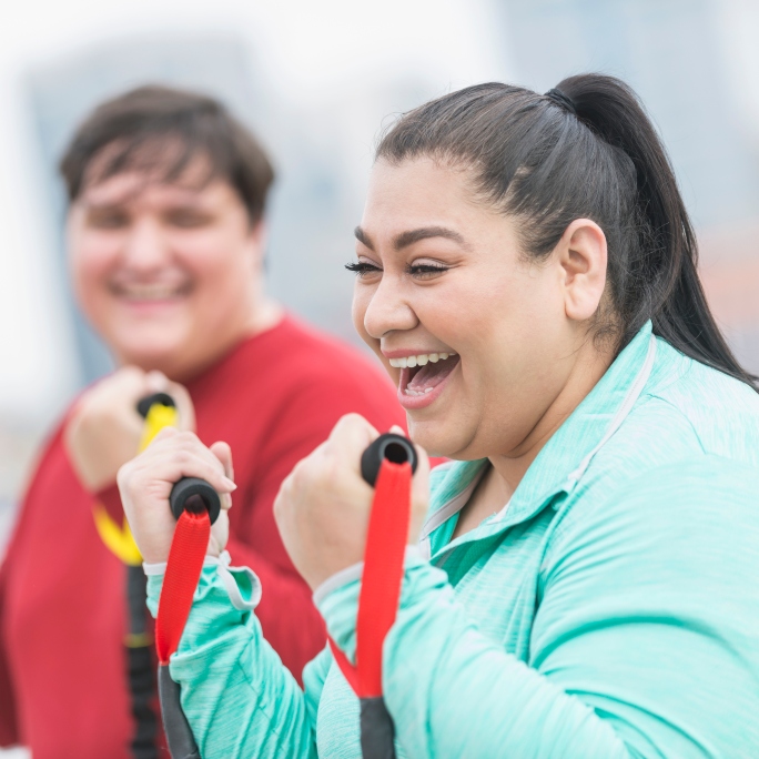 Woman exercising with resistant bands and smiling with workout partner in the background