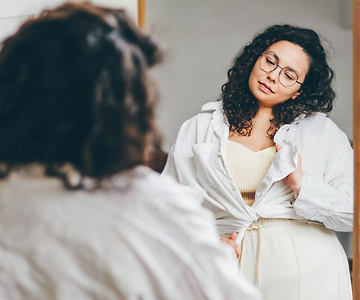Smiling woman looking in a mirror as she gets dressed in a white outfit.